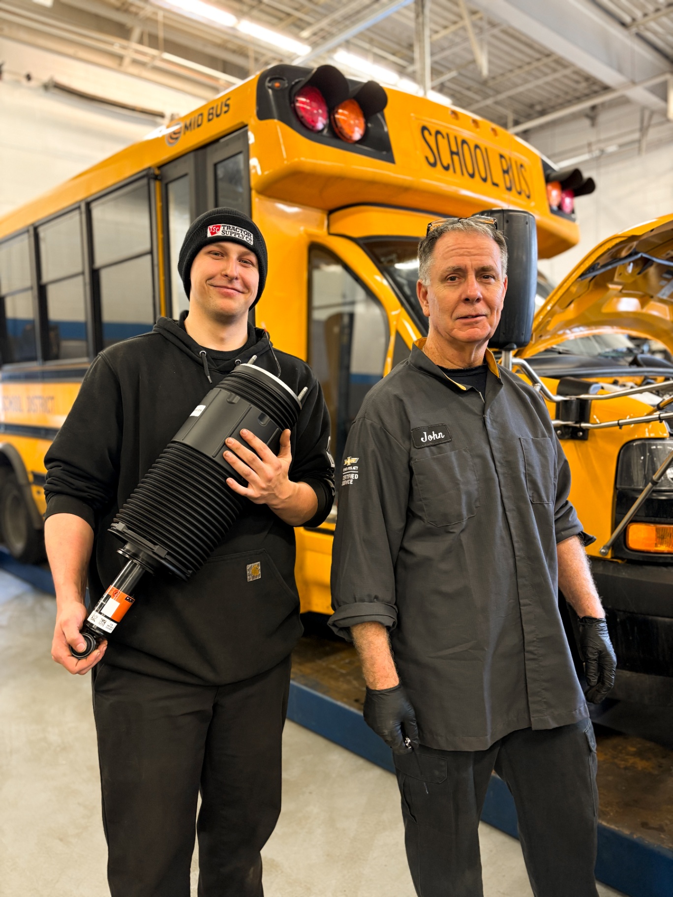 Two Patriot technicians in front of a school bus holding a replacement air strut